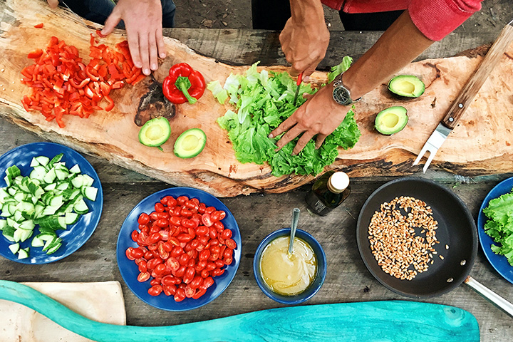 Fotografía cenital de dos pares de manos preparando frutas y verduras coloridas en una tabla de cortar de cocina