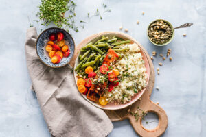 Fotografía cenital de un tazón de verduras y cereales saludables sobre una tabla de servir de madera con pequeños tazones de frutas, nueces y hierbas a su alrededor.
